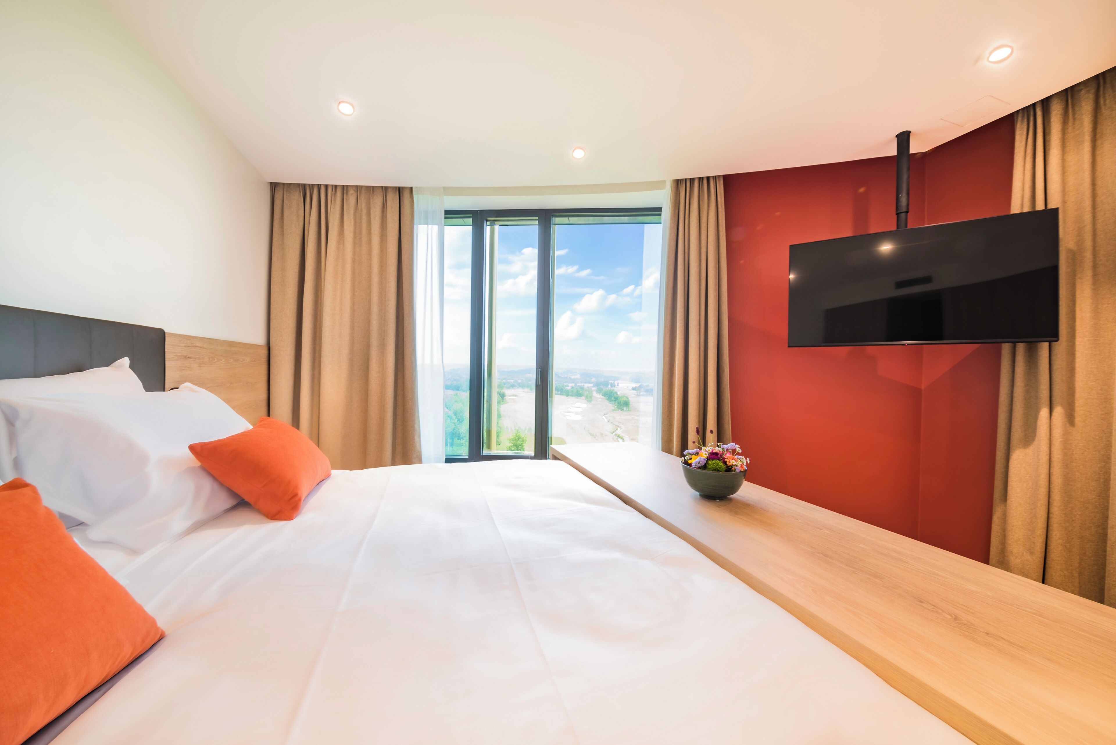 Ein Hotelzimmer mit Bett, Wandfernseher, einem Fenster mit Vorhängen und Blick ins Freie.