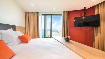 Ein Hotelzimmer mit Bett, Wandfernseher, einem Fenster mit Vorhängen und Blick ins Freie.