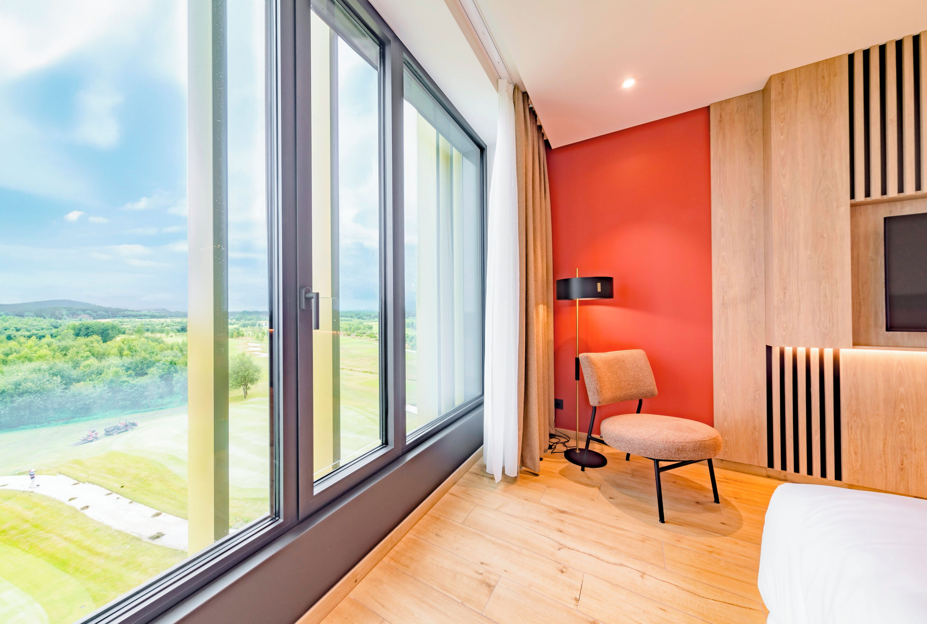 Habitación de hotel moderna con un ventanal grande, suelo de madera, pared de acento roja, una silla y una mesita de noche.