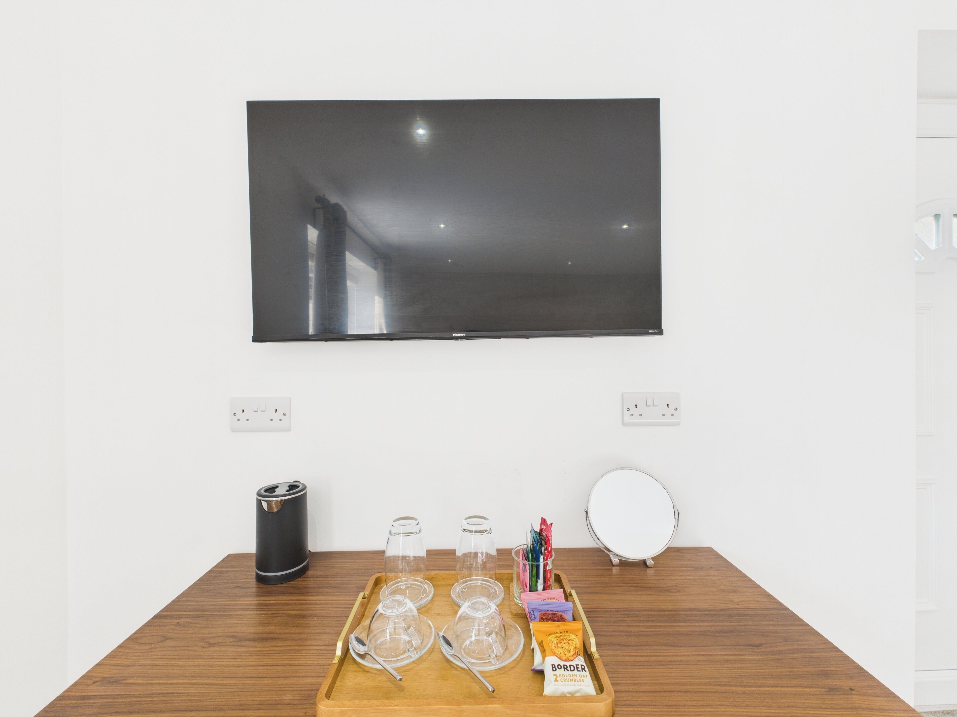 Une pièce avec un téléviseur fixé au mur, une table en bois sur laquelle se trouve un plateau avec des verres, une bouilloire et un autre plateau contenant des en-cas.