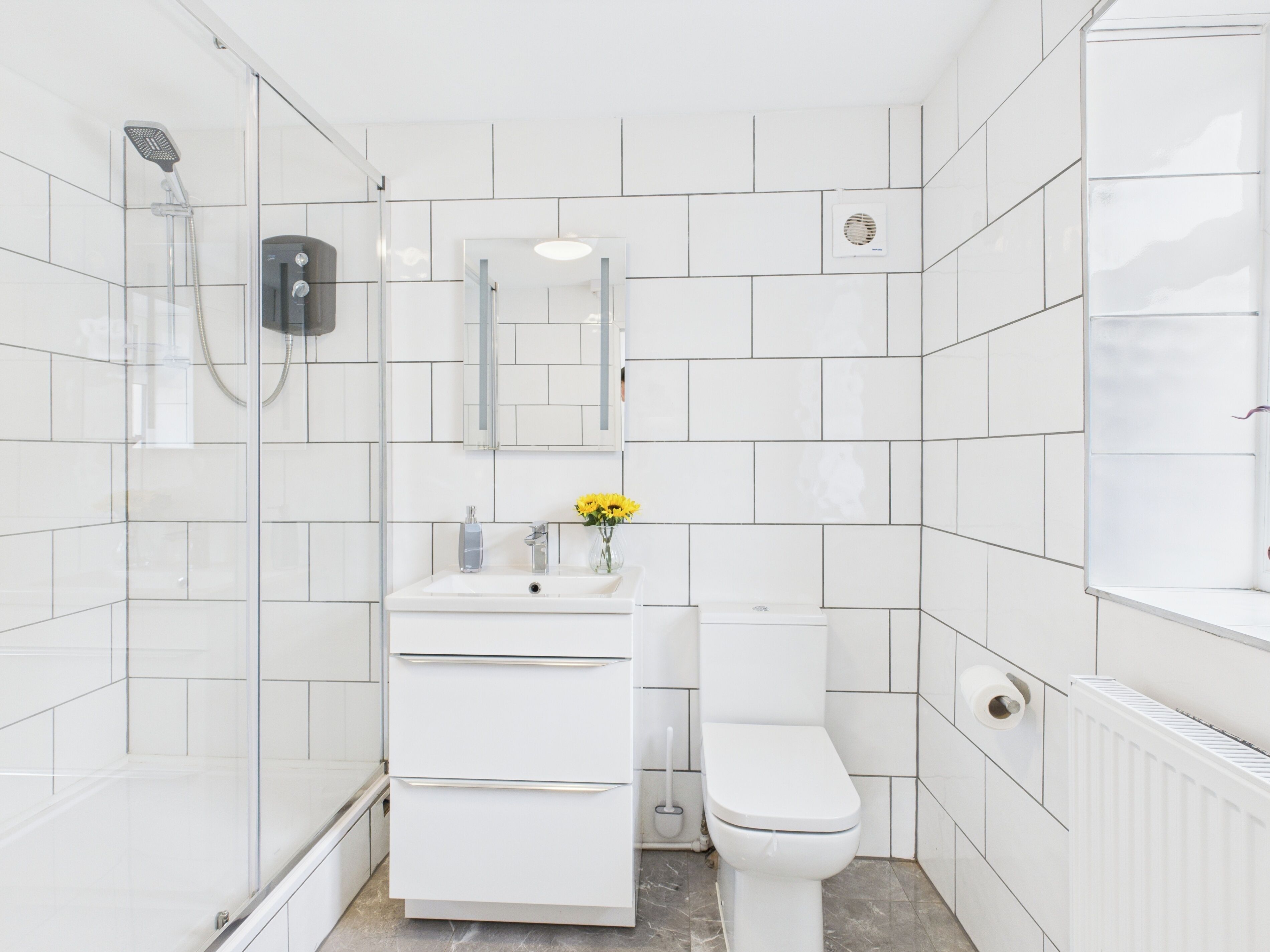 A modern bathroom with white tiles, a glass shower enclosure, a white sink with a mirror above it, and a toilet.