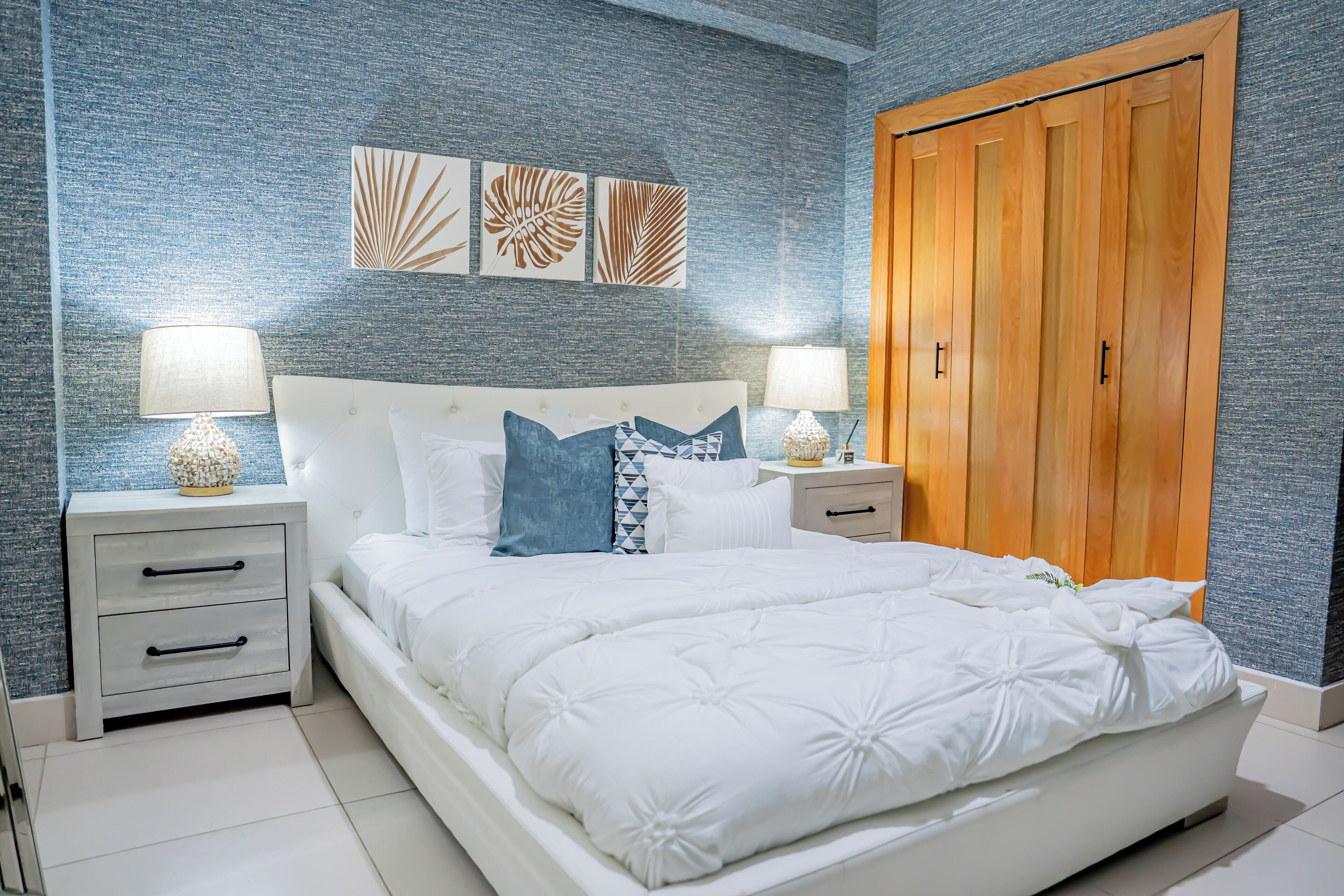 A bedroom with a large bed, two bedside tables with lamps, a wooden wardrobe, and decorative wall art.