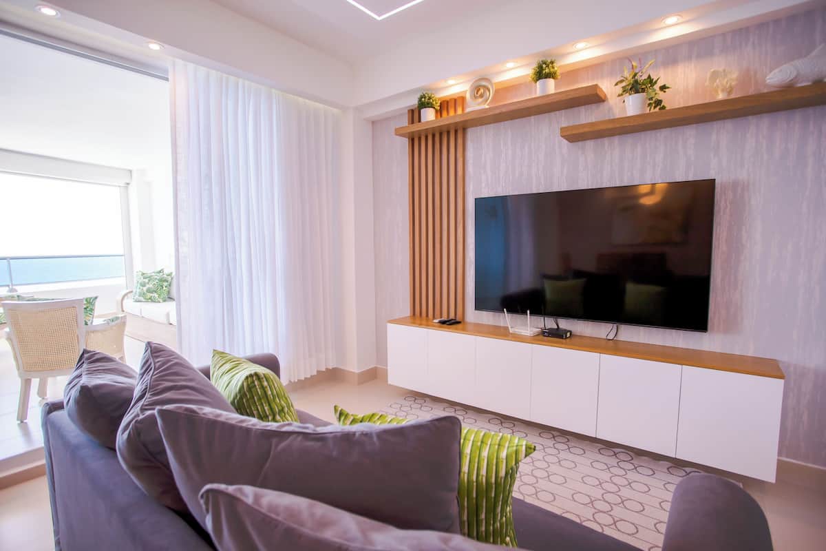 Ein modernes Wohnzimmer mit einem großen Flachbildfernseher, einem Sofa mit grünen und grauen Kissen und einem Holzregal mit Pflanzen.