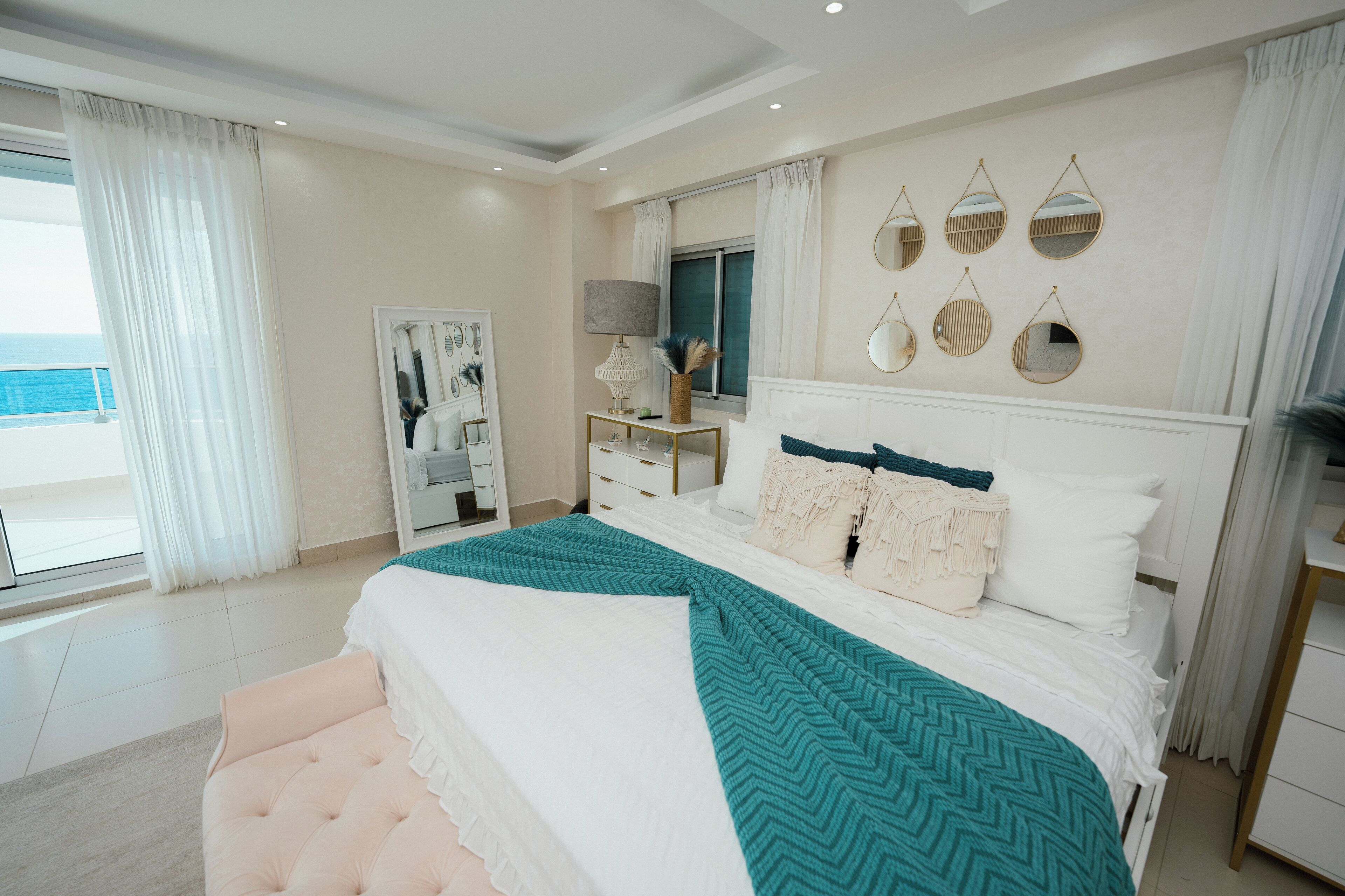 A bedroom with a large bed, a dresser, a vanity, and a view of the sea.