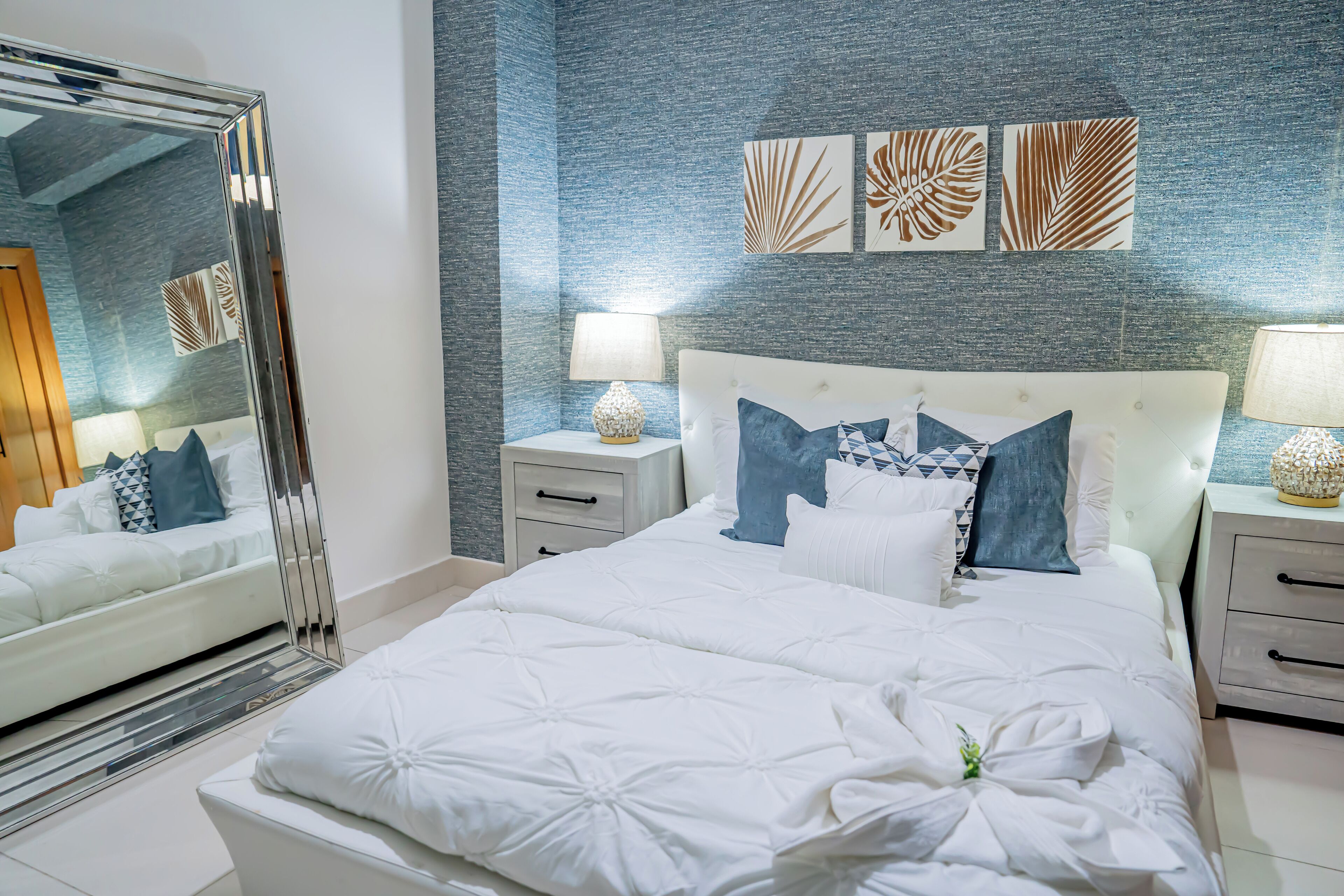Une chambre à coucher avec un grand lit, du linge de lit blanc, un miroir, des tables de chevet avec des lampes et des œuvres d’art murales.