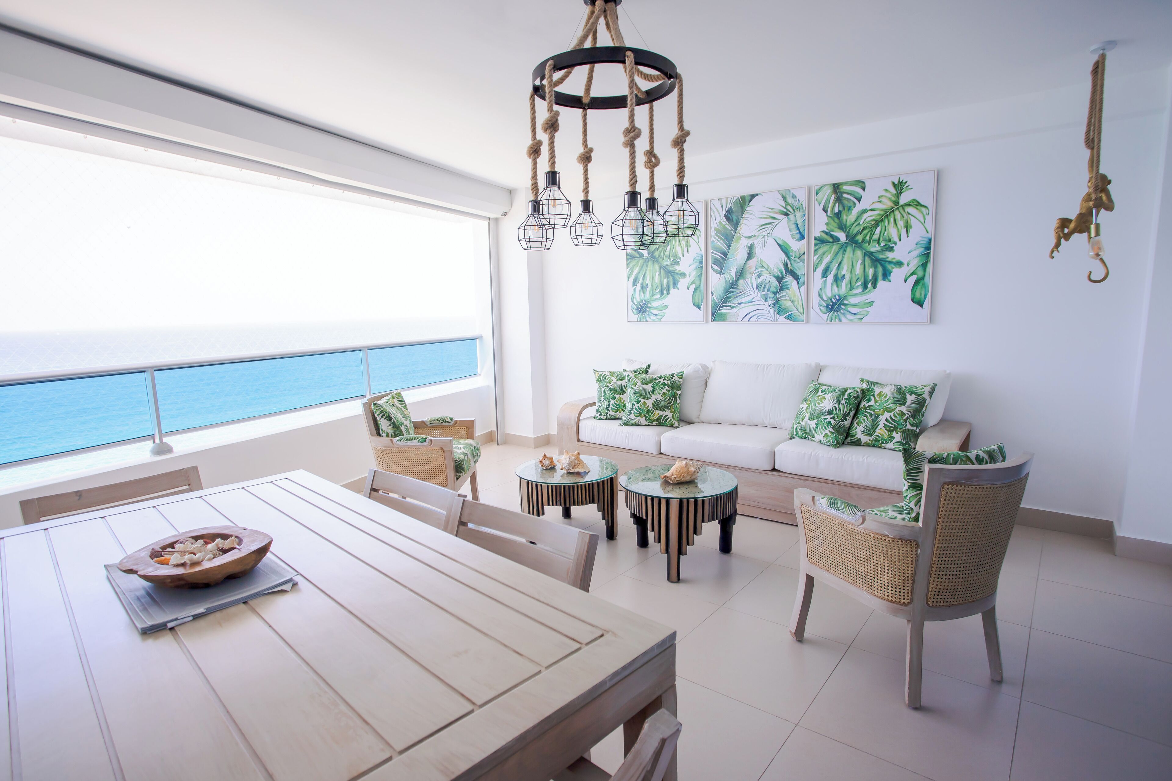 Un luminoso salón con vistas al océano, una mesa de comedor de madera, un sofá con cojines verdes y una lámpara original.