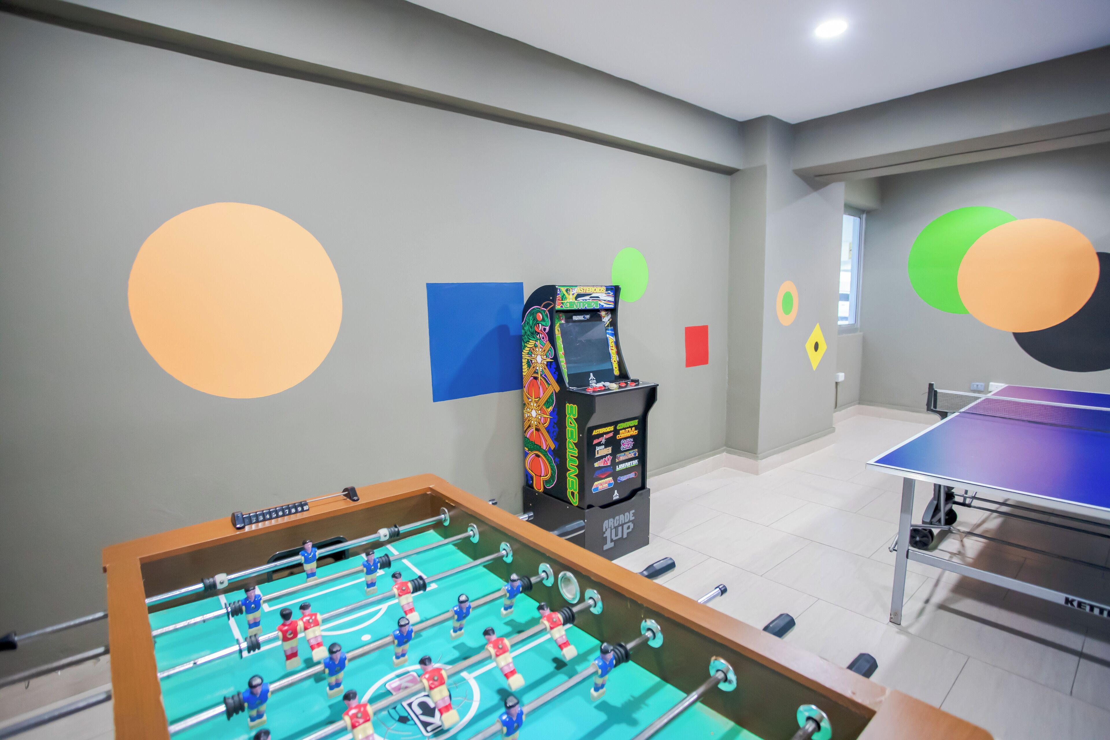 A room with a foosball table, a ping pong table, and a foosball machine.