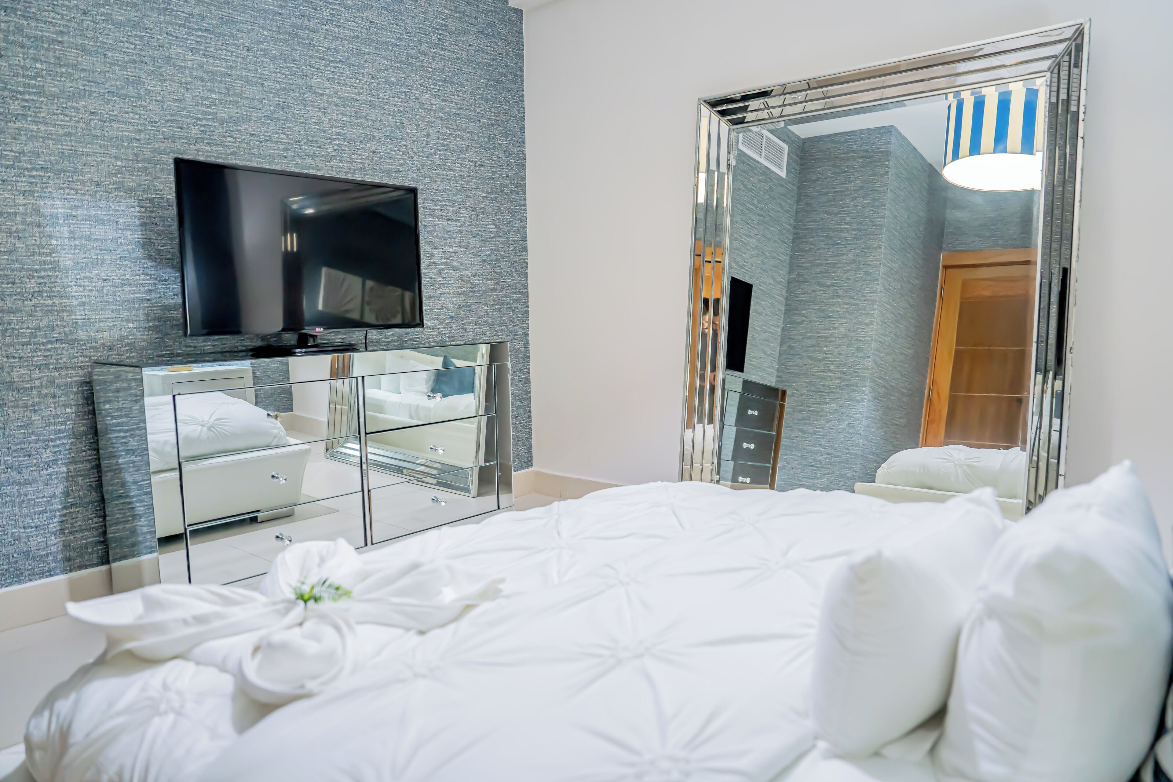 Quarto de hotel moderno com uma cama grande, uma TV de tela plana na parede, penteadeira de vidro e um espelho grande refletindo o ambiente.