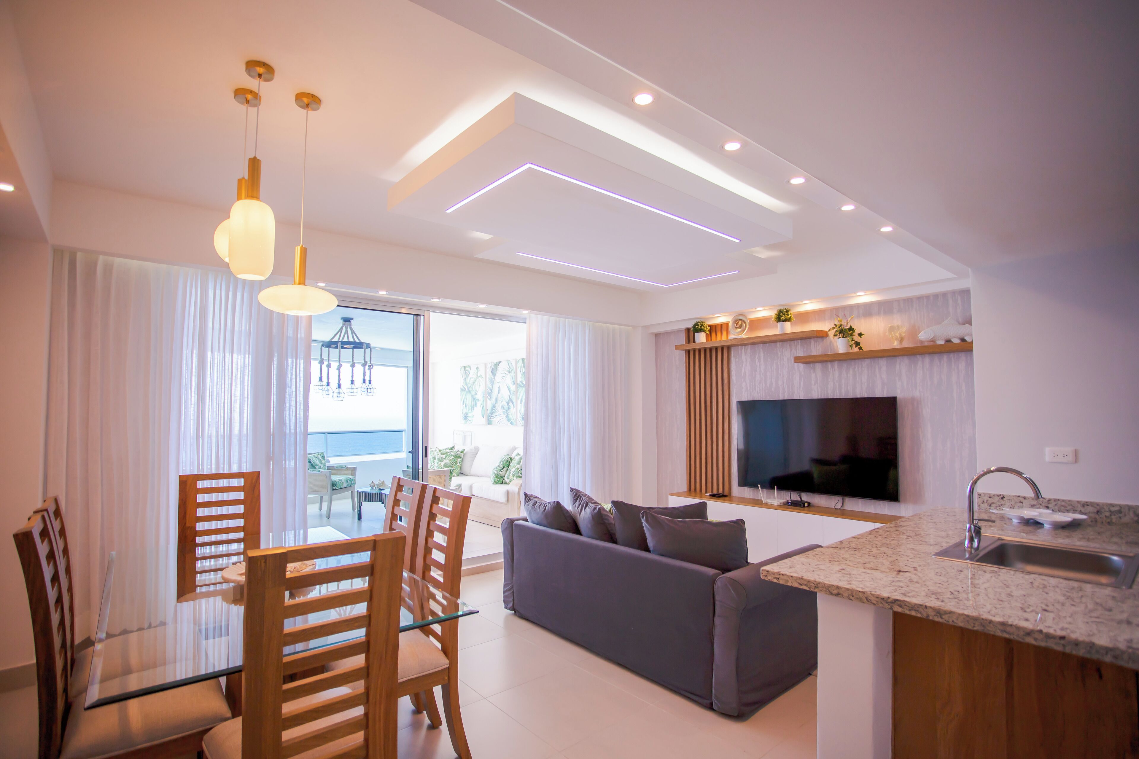 A modern living room with a grey sofa, wooden chairs, a glass dining table, and pendant lighting.