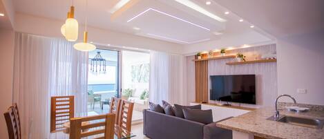 A modern living room with a grey sofa, wooden chairs, a glass dining table, and pendant lighting.