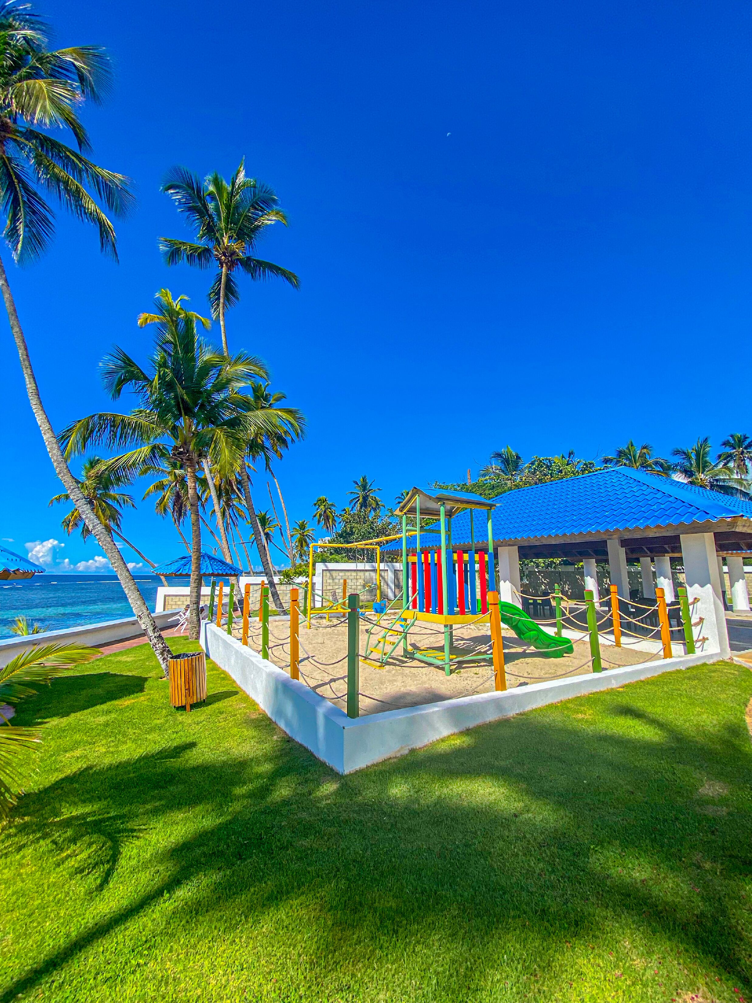 Un parque infantil con tobogán y estructuras para escalar, rodeado de palmeras y vista al mar.