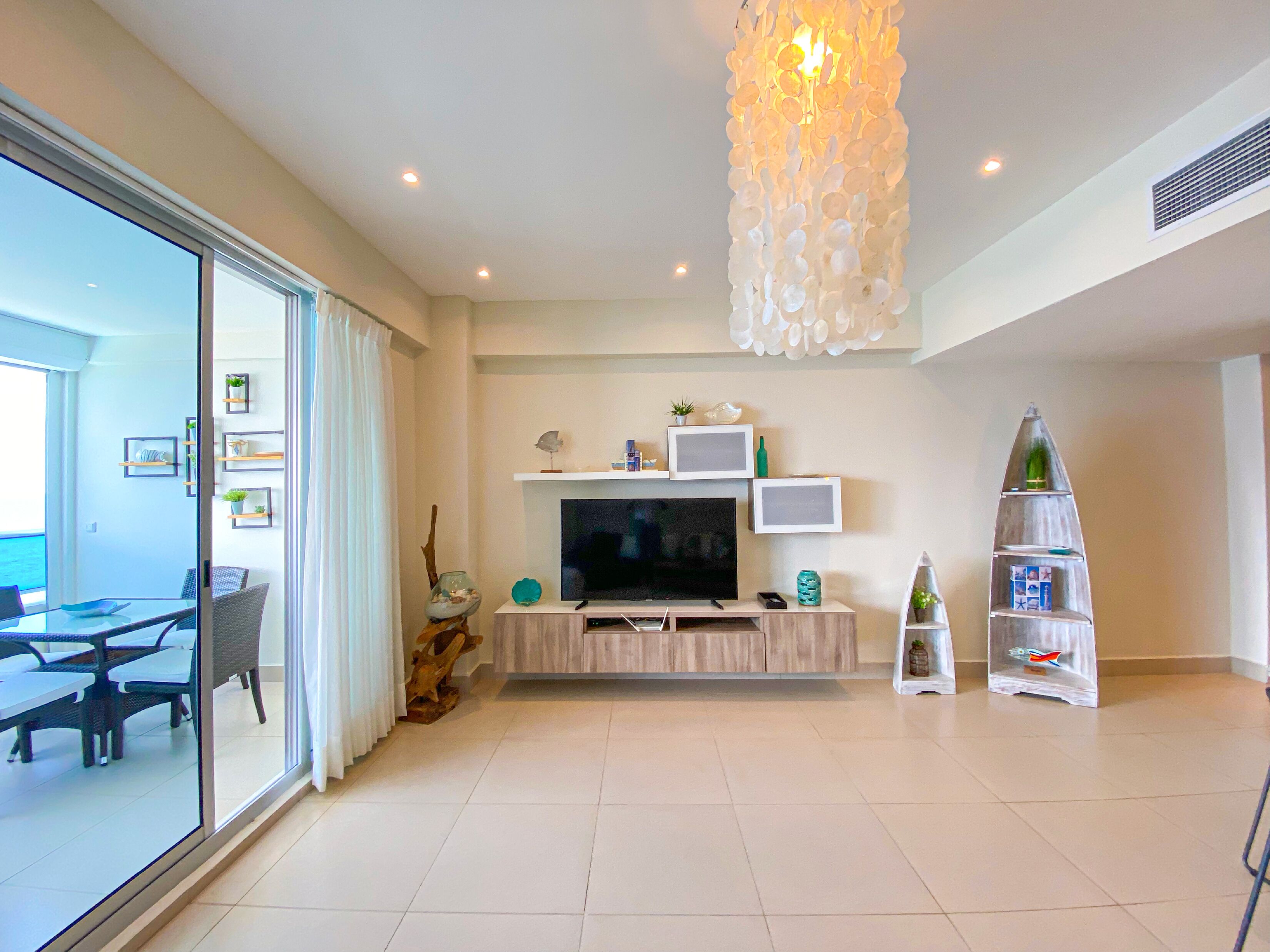 A modern living room with a large TV, a dining area with a glass table, and a balcony with a view.
