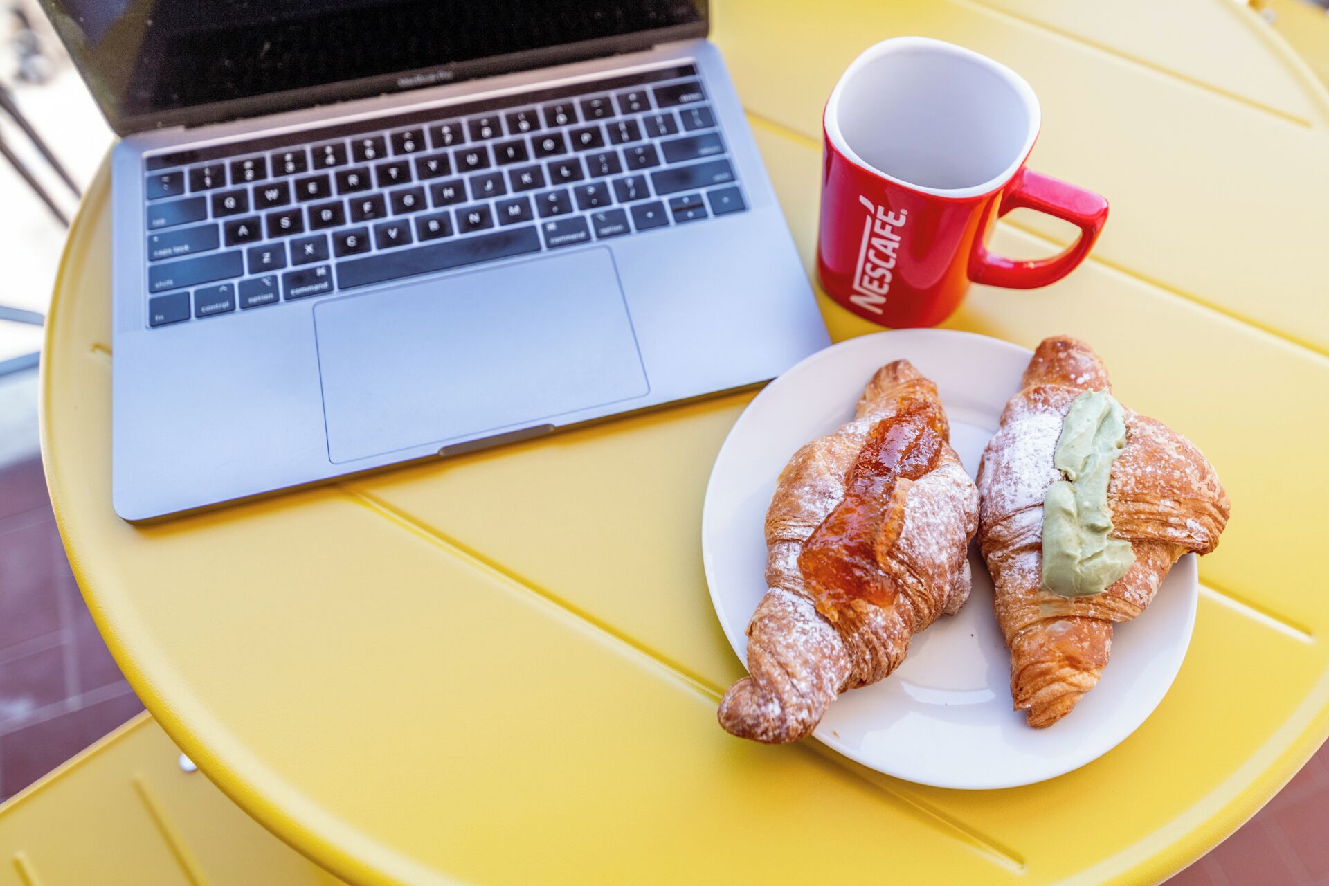A laptop on a yellow table with a red mug and two croissants on a white plate.