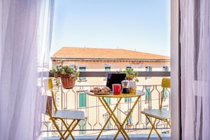 A balcony with a table, chairs, a potted plant, a coffee maker, and croissants.