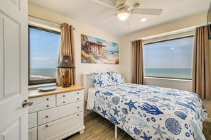 Un dormitorio con cama, mesita de noche, lámpara, cómoda y un cuadro de una escena costera.