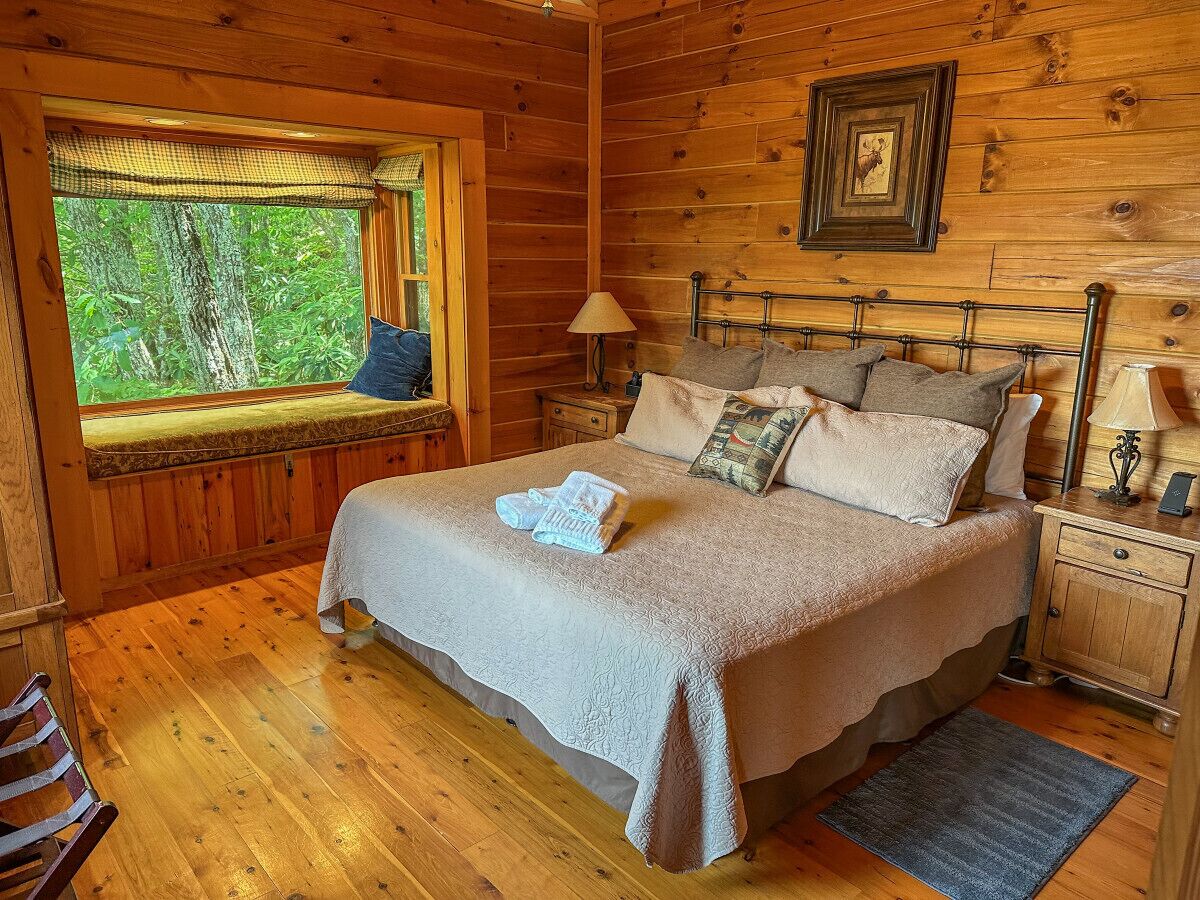 Une chambre dans une cabane en bois, avec un lit, une fenêtre donnant sur les arbres, une table de chevet avec une lampe et une petite table de nuit avec une autre lampe.
