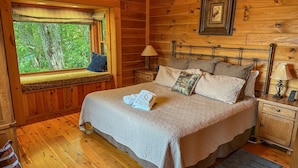 A wooden cabin bedroom with a bed, a window with a view of trees, a bedside table with a lamp, and a nightstand with a lamp. - Cloud 9-Log Cabin, Stunning View, Luxury Hot Tub, Gas Fire Pit, 30 min to Boone (Mountain City)