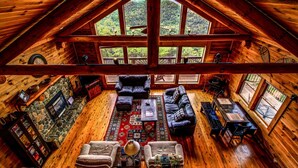 A spacious loft-style room with a stone fireplace, leather sofas, a dining table, and large windows offering a view of the forest. - Cloud 9-Log Cabin, Stunning View, Luxury Hot Tub, Gas Fire Pit, 30 min to Boone (Mountain City)