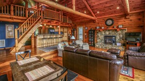 A spacious log cabin interior with a wooden staircase, a stone fireplace, leather seating, and a television. - Cloud 9-Log Cabin, Stunning View, Luxury Hot Tub, Gas Fire Pit, 30 min to Boone (Mountain City)