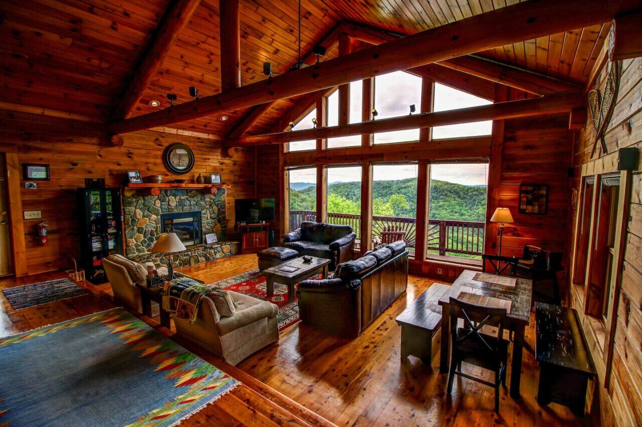 L’intérieur chaleureux d’un chalet en bois, avec une cheminée en pierre, des fauteuils en cuir et de grandes fenêtres offrant une vue sur les montagnes.