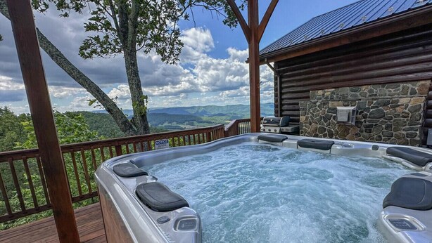 A hot tub on a wooden deck with a stone wall and a view of mountains. - Cloud 9-Log Cabin, Stunning View, Luxury Hot Tub, Gas Fire Pit, 30 min to Boone (Mountain City)