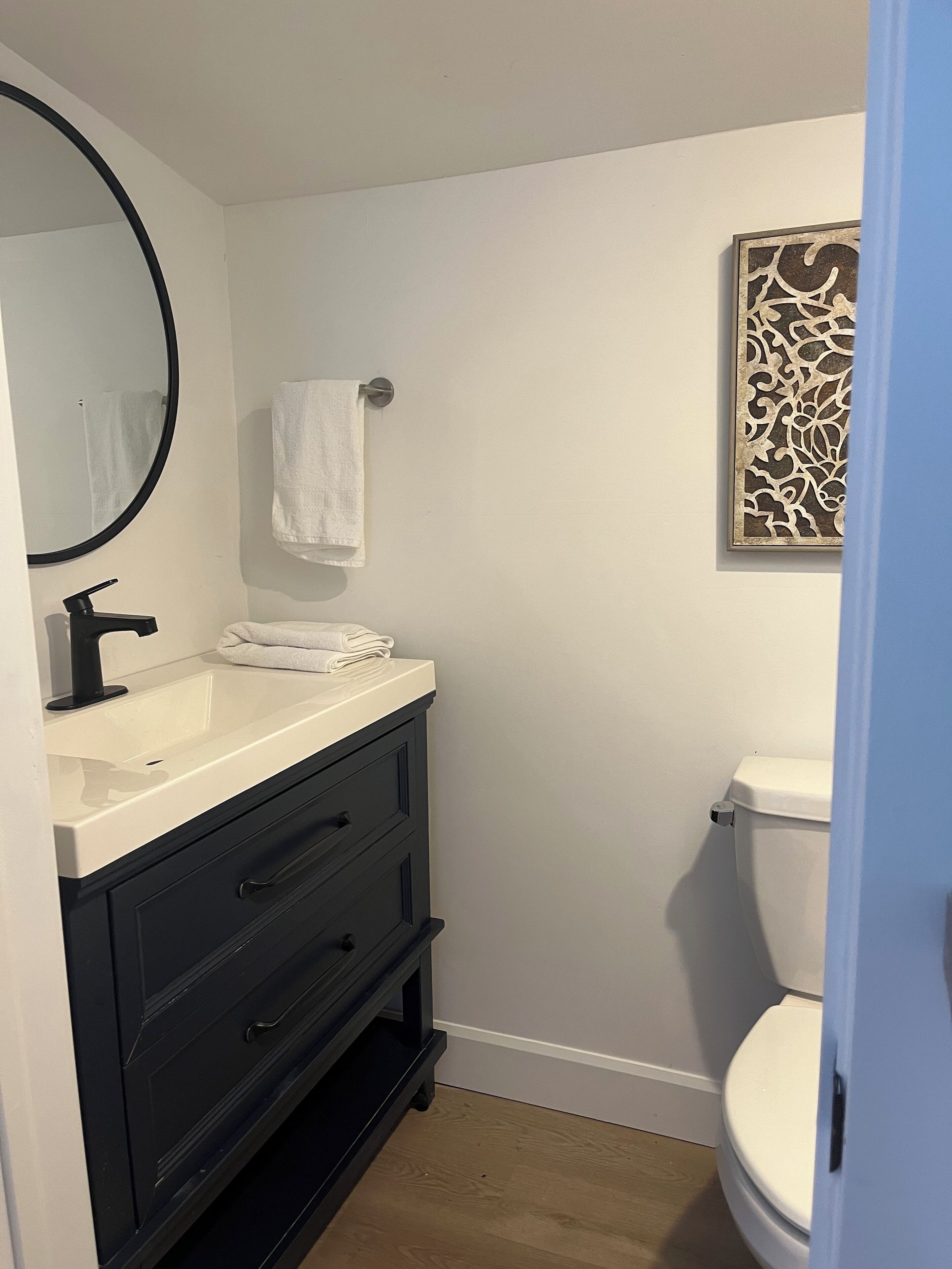 A bathroom with a white sink, black vanity, a round mirror, a white towel, a toilet, and a decorative wall piece.