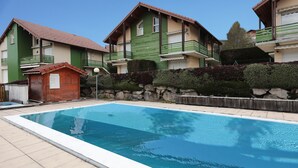 A swimming pool with a tiled deck, surrounded by houses with green and beige facades, stone walls, and a small wooden shed. - Domaine des Chênes Rouges - Appt 3 étoiles pour 4p dans chalet (Gérardmer)