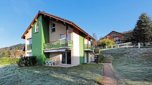 A two-story house with a green exterior and a brown roof, featuring a balcony and a small patio area with outdoor seating. - Domaine des Chênes Rouges - Appt 3 étoiles pour 4p dans chalet (Gérardmer)