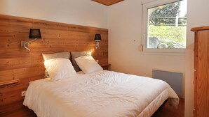 A bedroom with a wooden headboard, two bedside lamps, a bed with white bedding, and a window with a view of greenery. - Domaine des Chênes Rouges - Appt 3 étoiles pour 4p dans chalet (Gérardmer)