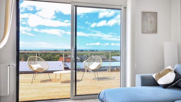 Ein Zimmer mit einer großen Schiebetür, die auf einen Balkon mit Blick ins Grüne führt. Auf dem Balkon befindet sich ein Bett mit blauer Bettwäsche und ein Sessel mit Kissen.