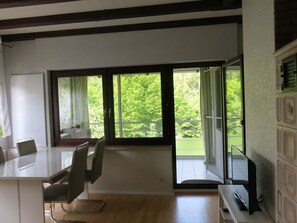 Ein modernes Zimmer mit einer großen Glasschiebetür, die auf einen Balkon führt, einem weißen Schreibtisch mit Stühlen und einem Fernseher auf einem Standfuß.