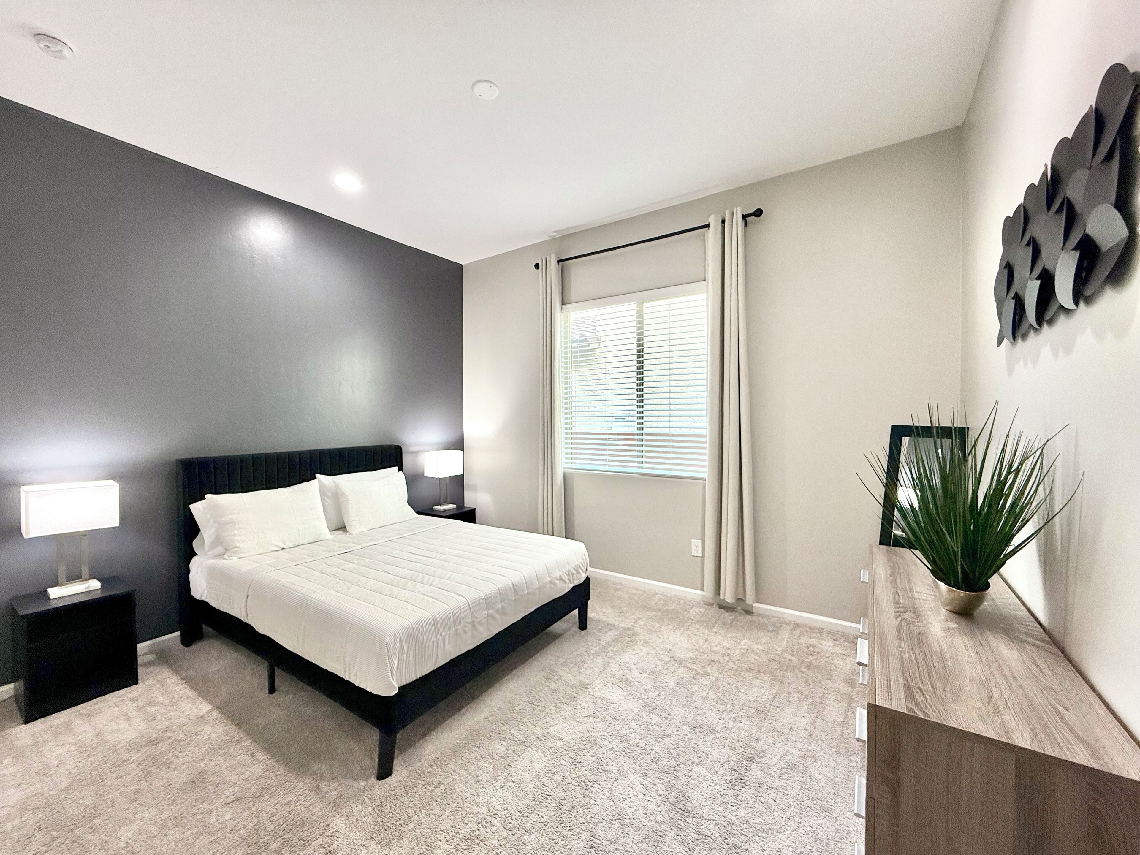 A modern bedroom with a large bed, bedside lamps, a window with blinds, and a plant on a wooden console.