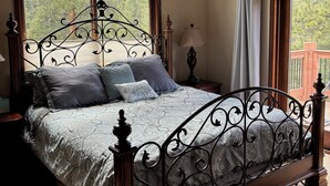 Ein Schlafzimmer mit einem großen Bett, einem Nachttisch, einer Lampe und Blick auf Bäume durch das Fenster.