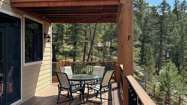 Eine Holzterrasse mit einem Glastisch und Stühlen, umgeben von einem Geländer und mit Blick auf eine bewaldete Landschaft.