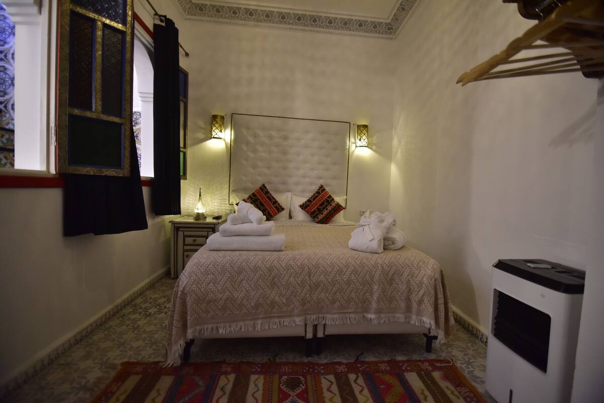 A bedroom with a bed, two bedside tables, a nightstand, a heater, and a window with curtains.