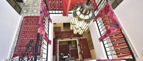 Una escalera de caracol con barandilla de estampado rojo y blanco, un candelabro colgante y vista de una habitación con alfombra estampada y una pequeña mesa.