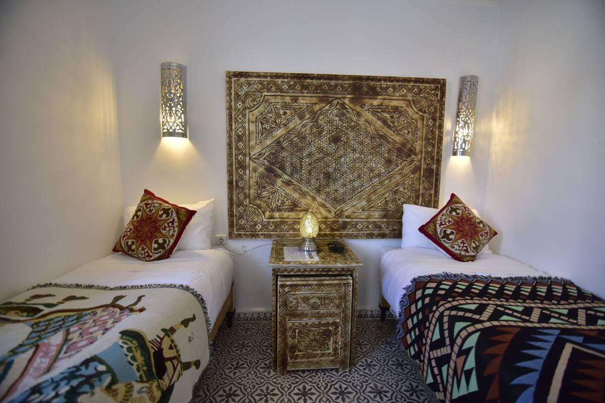 A room with two beds, patterned rugs, a decorative headboard, and wall-mounted lights.