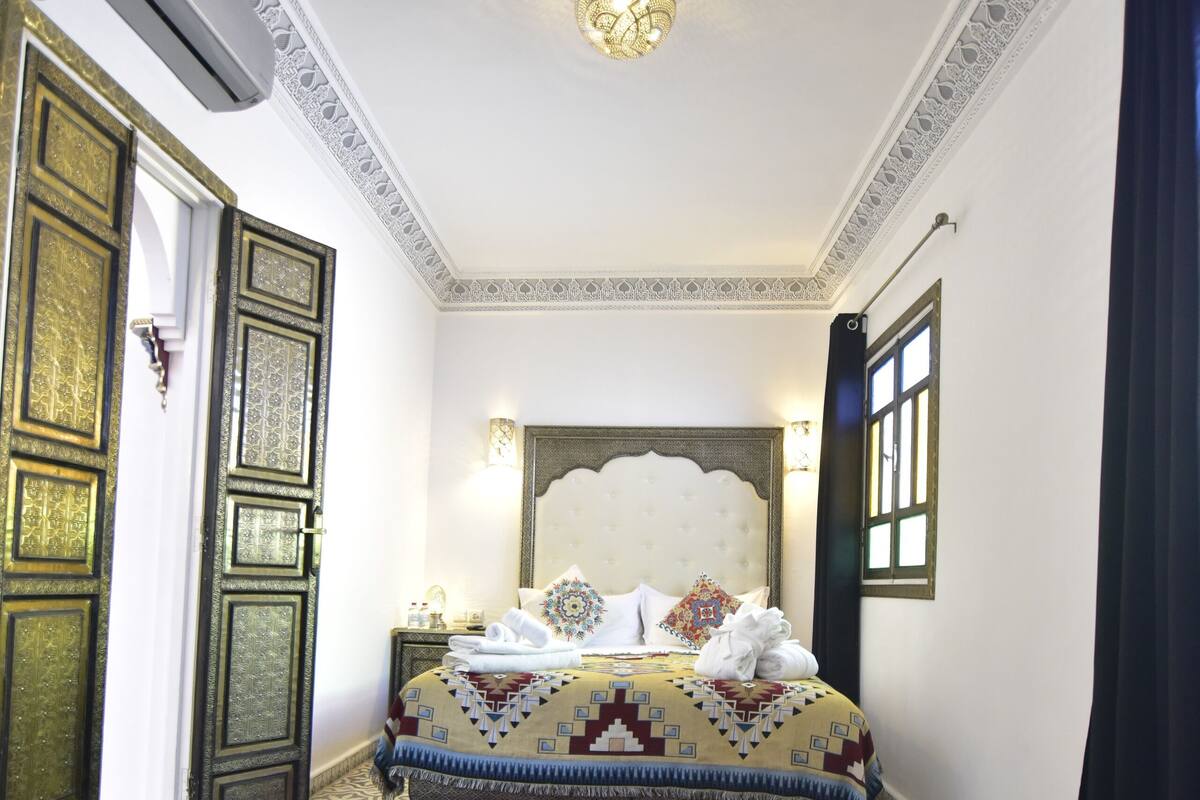 A room with a bed, patterned carpet, ornate ceiling, and a wall-mounted air conditioning unit.
