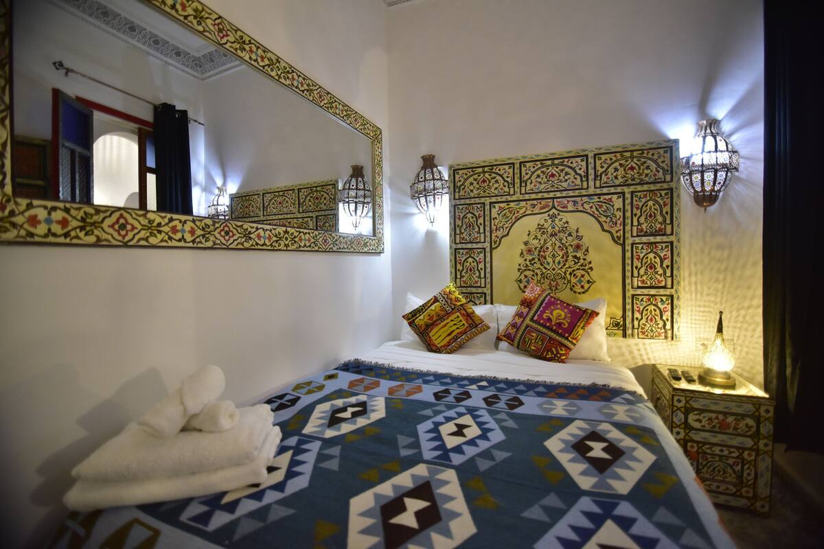 A hotel room with a large bed, a decorative headboard, a wall-mounted mirror, and a bedside table with a lamp.