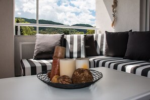 Et værelse med en sofa vendt mod et vindue med udsigt over et skovområde, et bord med en kurv nødder, og en dekorativ genstand på væggen.