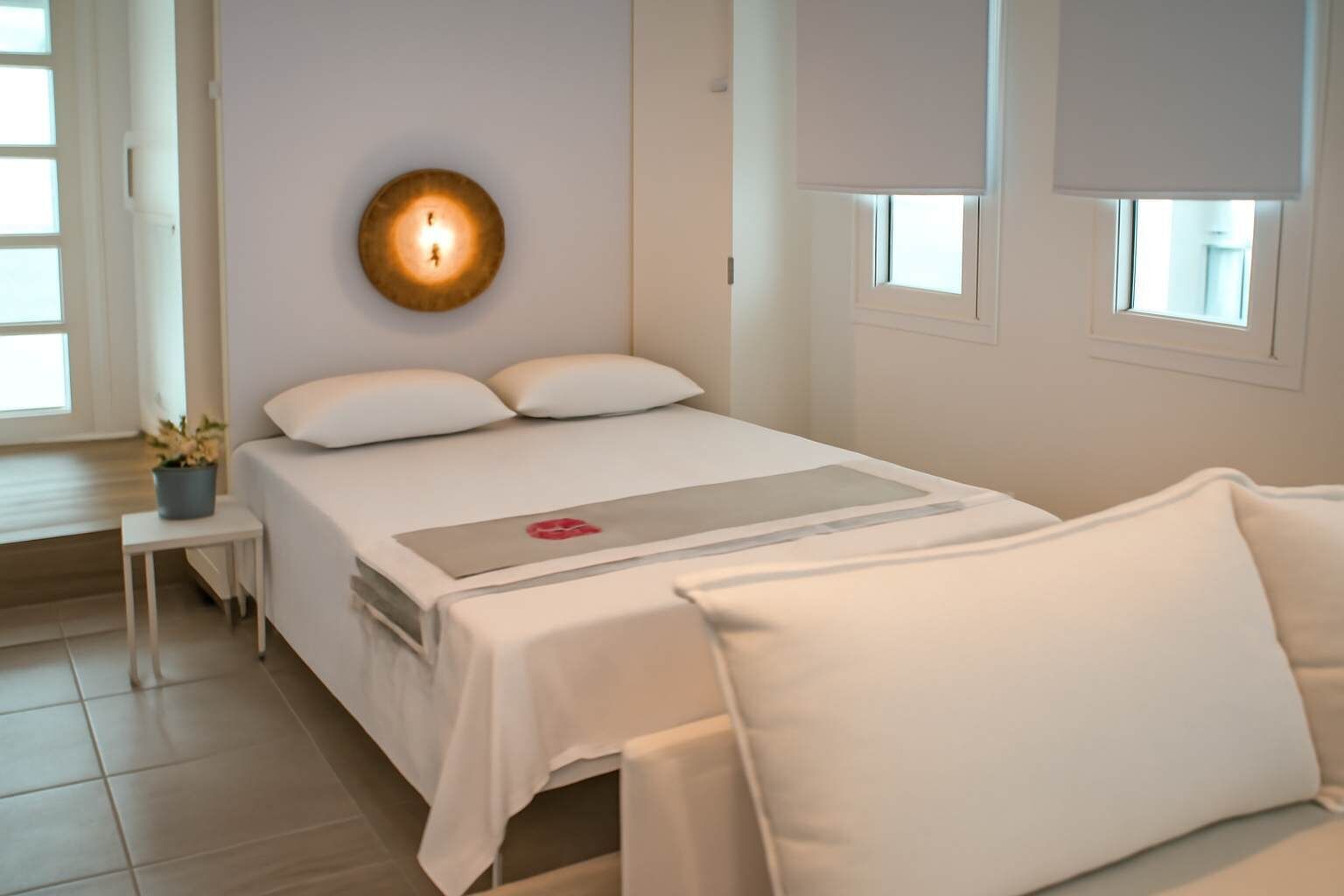 A hotel room with a bed, bedside table, wall-mounted light, and windows.