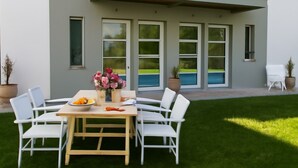 A modern house with a patio, white chairs, a table with flowers, and a potted plant.