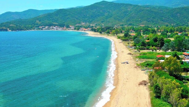 A coastal landscape with a beach, clear blue waters, and green hills in the background.