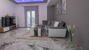 A modern living room with a grey sofa, a coffee table, a TV on a wooden stand, and a large window with curtains.