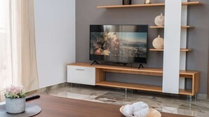 Moderni olohuone, jossa on litteänäyttöinen televisio, puuhyllyt, marmorilattia ja ruokapöytä, jonka päällä on koristeellisia lautasia.