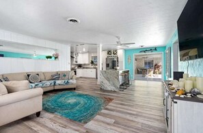 Un salón amplio con un sofá color crema, un tapete verde azulado y una cocina con armarios blancos y una isla de mármol.