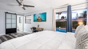 A bedroom with a bed, a desk, a ceiling fan, a painting of a turtle, and a view of a pool and palm trees. - Pool Oasis 2 Blocks from Beach (Port Orange)