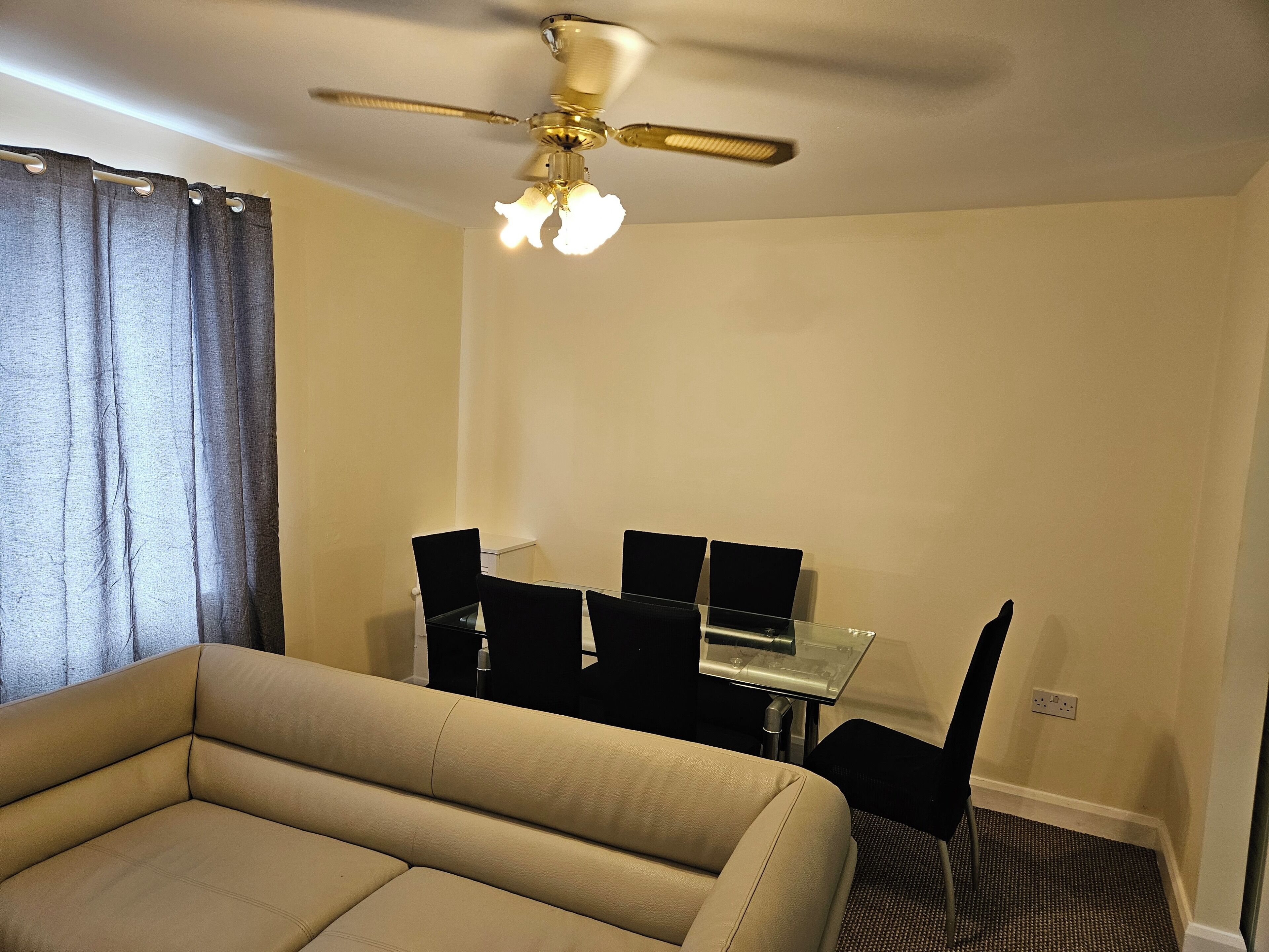 A room with a beige sofa, a dining table with chairs, a ceiling fan, and a window with curtains.