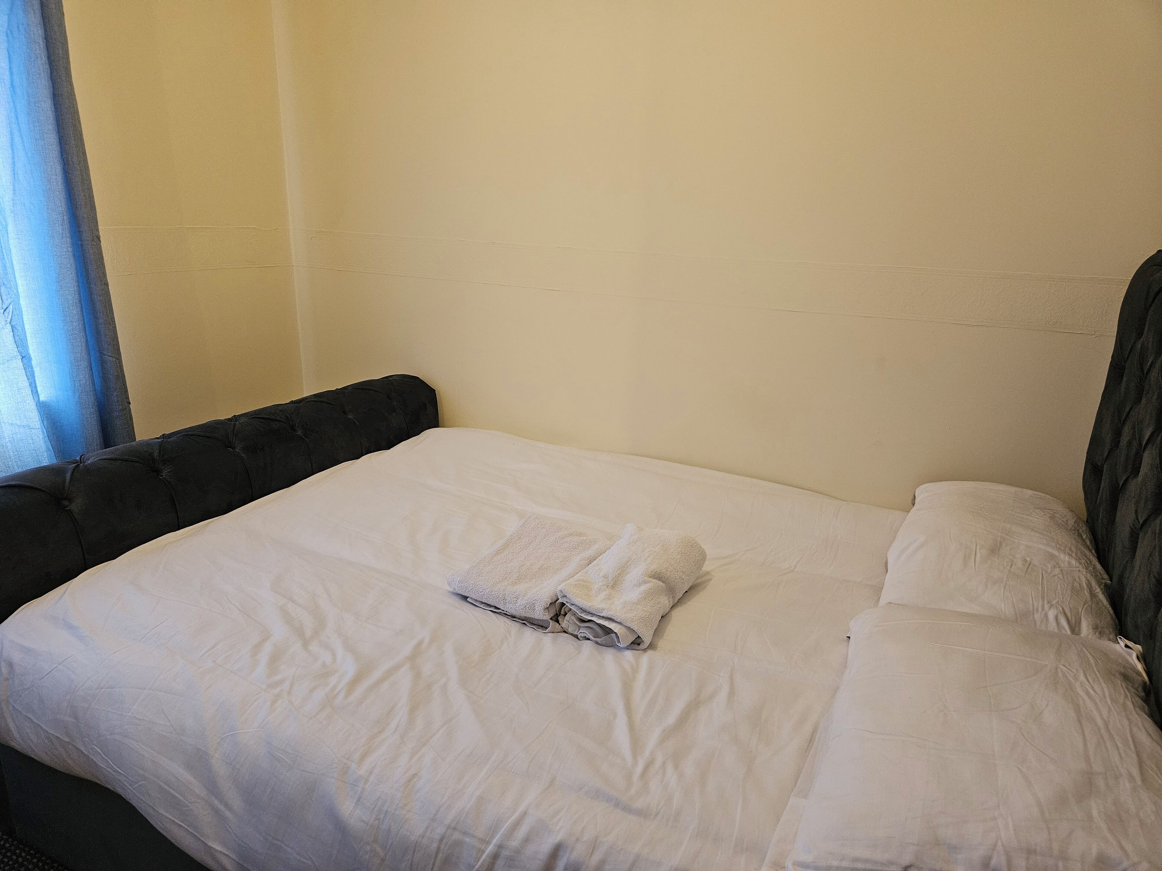 Una cama bien hecha con sábanas blancas y una toalla doblada, cabecera oscura y pared lisa.