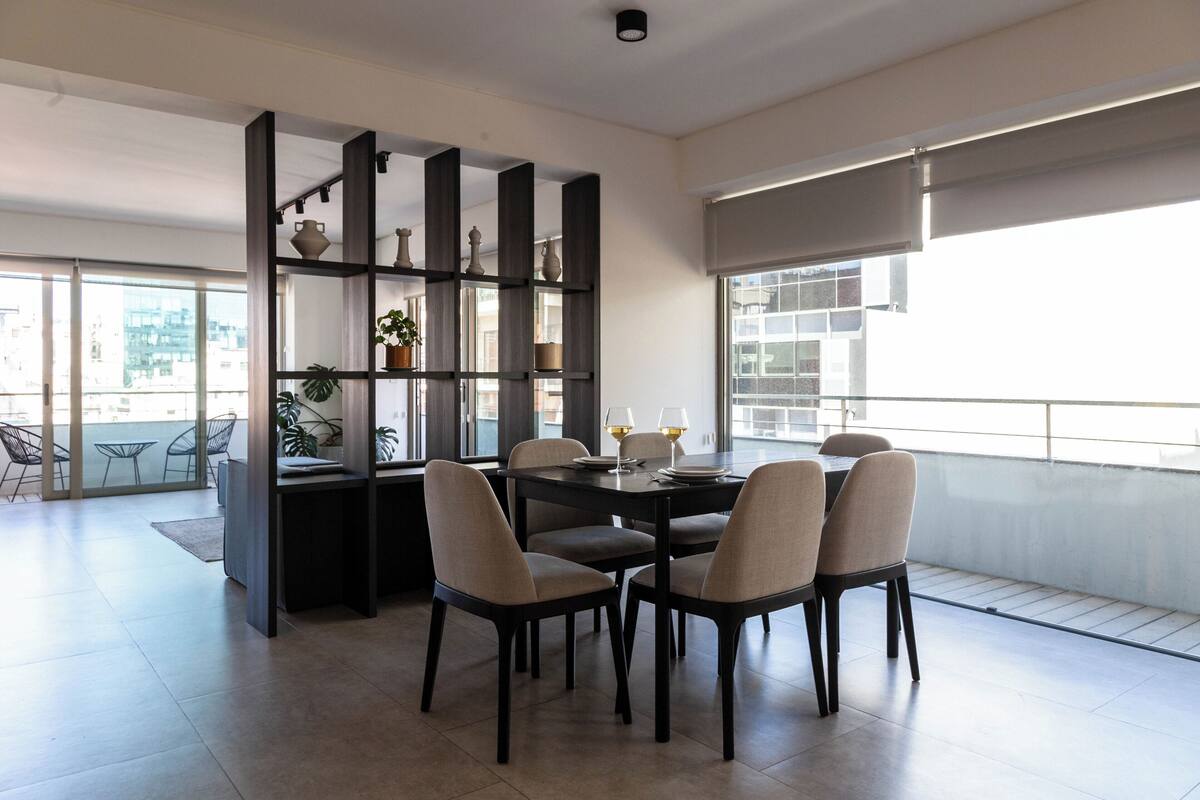 A modern dining area with a dark wooden table and chairs, a bookshelf with decorative items, and large windows offering a view of the city.